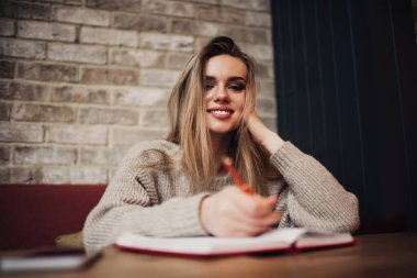 Örgü örülü süveter giymiş neşeli genç bir bayan rahat bir çatı katında oturuyor ve gülümseyip kameraya bakarken notlar alıyor.