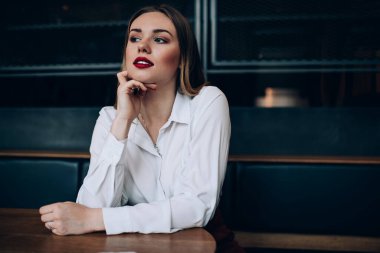 Beyaz gömlekli, rüya gibi bir kadın. Deri koltukta, tahta masada oturuyor ve başka tarafa bakıyor.