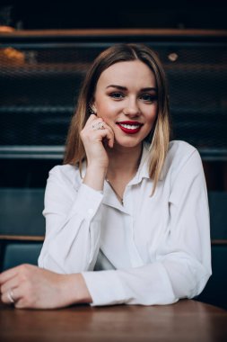 Resmi giyinmiş neşeli genç bir kadın kırmızı dudaklı, elleri çenesinde ve dişleri üzerinde oturmuş kameraya bakarken gülümsüyor.