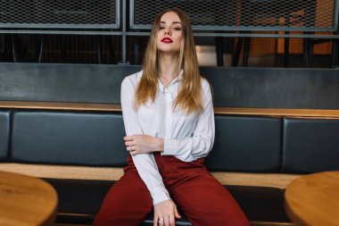 Modern restorandaki ahşap masaların arasında oturmuş kameraya bakarken modaya uygun giysiler içinde kendine güvenen genç bir kadın.