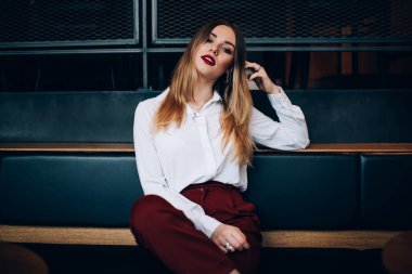 Kırmızı dudaklı, ciddi bir genç bayan deri bir bankta dinleniyor ve kameraya bakarken kafasına dokunuyor.