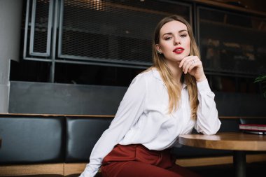 Modern kafede oturmuş kameraya bakarken şık elbiseli, iyimser genç bir bayan.