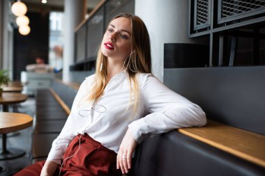 Kafedeki kanepede dinlenen ve gün ışığında müzik dinleyen sarışın genç bir kadının yan görüntüsü.