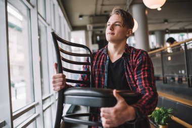 Kafede siyah deri sandalyeyle gezen, gülümseyen ve pencereden dışarı bakan, günlük kıyafetli, şık, genç bir erkek.