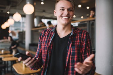 Modern kafe molasında beyaz sütunun önünde dururken cep telefonuyla gülümseyen rahat elbiseli neşeli genç adam.