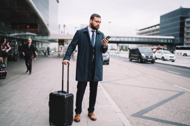 Bavulları havaalanı terminalinin yanında duran ve akıllı telefonları tararken araba bekleyen resmi giysili tam boy erkek.