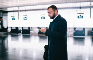 Havaalanı koridorunda dikilip uçuş beklerken cep telefonuyla sörf yapan siyah ceketli ciddi bir erkek işçinin yan görüntüsü.