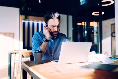 Düşünceli, sakin, zeki, sakallı, gri saçlı erkek. Ofisteki dağınık masada laptopla çalışırken telefonla konuşuyor.