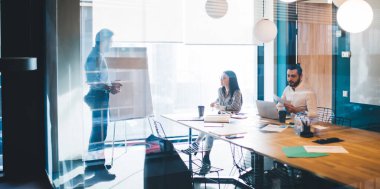 Koyu saçlı kadının yanında oturan sakallı genç adam ve bilgisayarını kullanarak çağdaş tasarım bürosunda projeyi sunuyor.