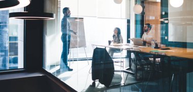 Çağdaş tasarım işyerindeki ofis malzemeleriyle masada oturan genç iş arkadaşlarıyla yan yana duran iş adamı.