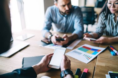 Ofis malzemeleriyle masada otururken elinde boş ekranlı akıllı telefon tutan ve akıllı telefonları olan iş arkadaşlarının yanında doküman taşıyan bir adam.