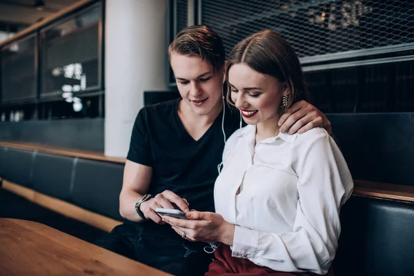 Pozitif genç trend giyinmiş kadın ve erkek akıllı telefon kullanırken kucaklaşıp gülümsüyor ve müzik listesinden müzik dinliyor.