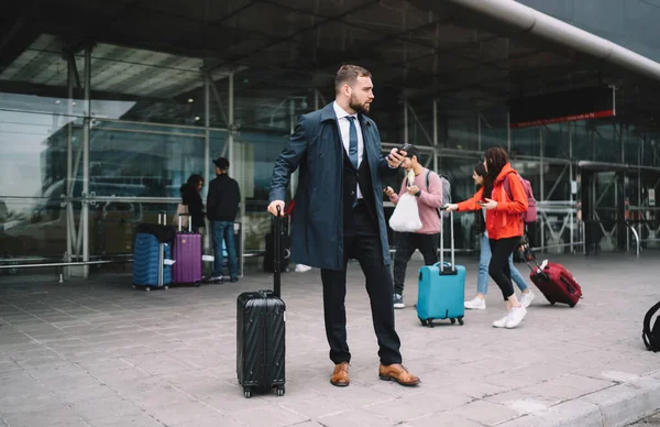 空港ターミナルに立つスーツケースを着て スマートフォンを使用しながら輸送を待っていると離れて見て正式な服装で自信のあるビジネスマンの完全な体 — ストック写真
