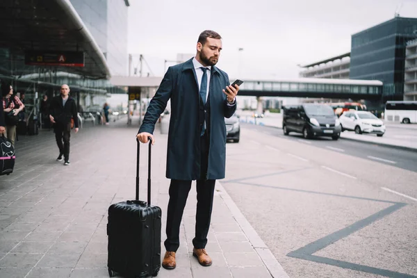 空港ターミナルの近くに荷物を置き スマートフォンを閲覧しながら車を待っている正式な摩耗で濃縮男性の完全な長さ — ストック写真