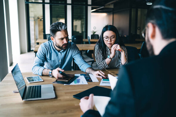Businessman pointing at document and explaining idea to female colleague while sitting at table and discussing results of marketing research