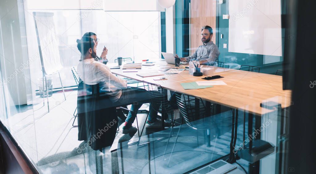 Group of serious colleagues sitting at table with laptop and documents and discussing details of business project while having meeting in conference room