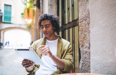 Gündelik kıyafet giyen ciddi etnik erkek öğrenci gündüz projesi üzerinde çalışırken sokak kafesinde kitap okuyor ve tablet kullanıyor.