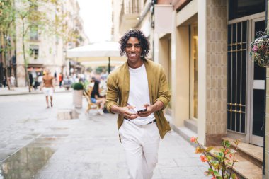 Modern binaların yakınında gezinirken ve kameraya bakarken akıllı telefon kullanarak gülümseyen sıradan giyinmiş neşeli, genç bir Afrikalı Amerikalı yaya.