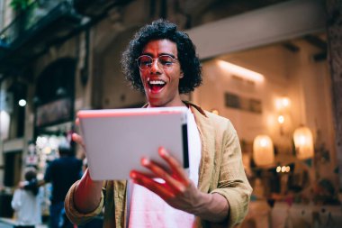 Şehir caddesinde terasta dikilirken tablet kullanan gündelik gözlüklü, mutlu etnik erkek.