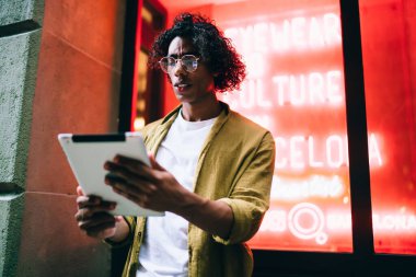 Düşük açılı, günlük giysiler ve gözlüklü etnik erkek şehir caddesinde duruyor ve tablet üzerinde internette geziniyor.