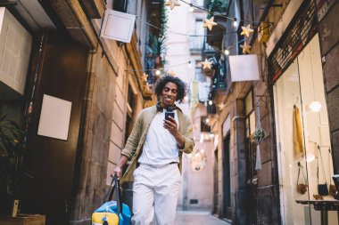 Dar geçitte yürürken cep telefonunda gülümseyen ve mesajlaşan el çantalı neşeli etnik adamın alçak açısı.