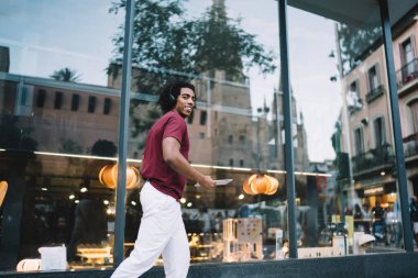 Gündelik giysiler içinde gülümseyen genç bir erkek şehir caddesinde yürüyor ve kulaklıkla müzik dinliyor ve kameraya bakıyor.