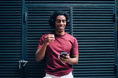 Gülümseyen etnik erkek, sağ eliyle kaldırdı kulaklıkla müzik dinledi ve metal çubuk kapısına yaslandı ve ekrana baktı.