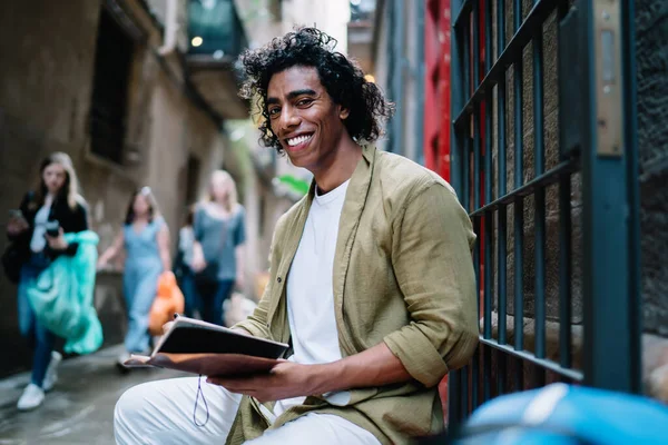 Side view of positive young black male student in trendy clothes smiling and reading notebook while relaxing on porch of old building and looking at camera