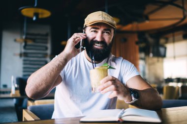 Sakallı erkek, kafede oturmuş soğuk bir şeyler içiyor. Kameraya bakmadan önce cep telefonuyla konuşuyor.