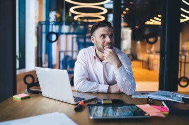 Neşeli sakallı iş adamı resmi kıyafetler giyer. Masada oturup güncel ofiste çalışırken gülümser ve dalgın dalgın dalgın bakar.