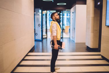 Asansörü beklerken elinde belgelerle salonda duran kulaklıklı modern erkek işçinin yan görüntüsü.