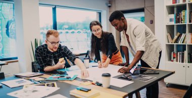 Genç, ciddi, çok ırklı iş arkadaşları gündelik giysiler içinde kahve fincanları ve ofis arşivleri ile masaya planlar çiziyorlar. 