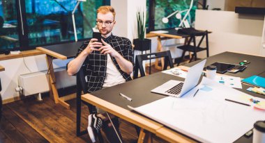 Genç hippi, dizayn projesi için saatlerce akıllı telefon mesajlarını kontrol ediyor dizüstü bilgisayarında dizüstü bilgisayarla oturuyor ve tasarım taslağı çiziyor, Kafkas milenyum adamı cep telefonunda sohbet ediyor.