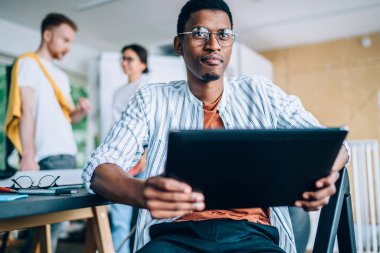 Düşünceli Afrikalı Amerikalı bir erkeğin günlük giysileri ve gözlükleriyle tablet kullanarak serbest çalışma projesini görmezden gelmesi.