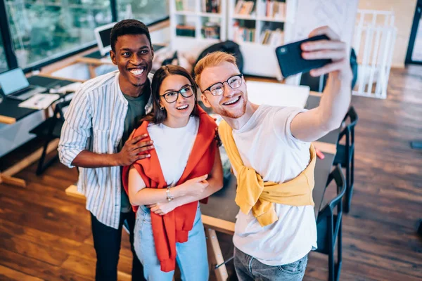 From above happy diverse people in casual clothes cheerfully smiling and using smartphone to take selfie while standing near table in office