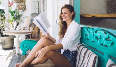 Koyu renk kıvırcık saçlı, beyaz tişörtlü, kot şortlu, renkli bankta oturmuş kitap okuyan mutlu bir kadın.