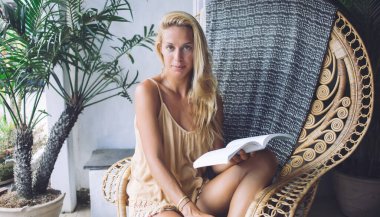 Çekici sarışın kadın, dizleri bükülmüş bir şekilde kapıdaki hasır koltukta yeşil bitkinin yanında oturuyor ve açık bir kitap tutuyor.
