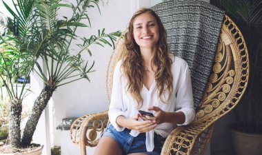 Güzel bir kadın güneşli bir günü terasta hasır sandalyede oturup gülerek ve cep telefonunda komik mesajlar okuyarak geçiriyor.