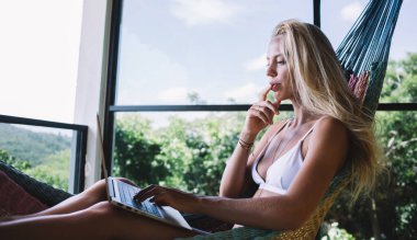 Beyaz bikinili, rahat bir hamakta dinlenen ve terasta dizüstü bilgisayar kullanan sakin, bronzlaşmış bir kadının yan görüntüsü.