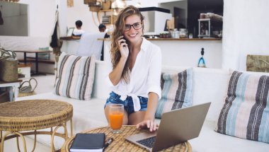 Telefonda konuşan çekici bir kadın iş sırasında kameraya bakıyor. Modern yazlık kafedeki sepet sehpasında laptopta.