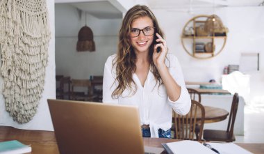 Gözlüklü, gülümseyen yetişkin bir yönetici kameraya bakıyor ve telefon görüşmesi yapıyor. Bu sırada kafeteryada planlamacı ve dizüstü bilgisayarla birlikte duruyor. Bulanık arka planda.