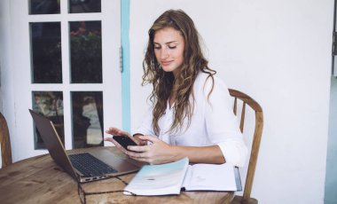 Kıvırcık saçlı, serbest çalışan, dizüstü bilgisayarını karıştıran, kitap okuyan ve yaz sıcacık verandasında, ahşap masada cep telefonuyla mesajlaşan bir kadın.