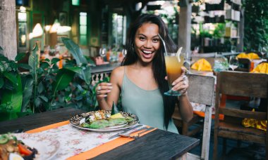 Neşeli, genç, etnik kadın vejetaryen günlük kıyafetlerle bir bardak taze narenciye ile kadeh kaldırırken tropikal kafe terasında sağlıklı bir akşam yemeği yiyor.