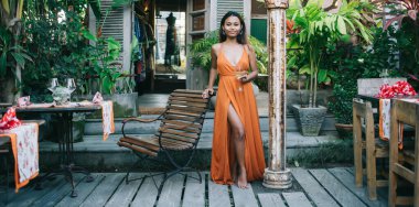 Süslü, maksimum kesim elbisesi içinde, egzotik kafenin terasında bir bardak şarapla dikilirken ahşap sandalyeye yaslanmış muhteşem, genç, etnik esmer bir kadın.