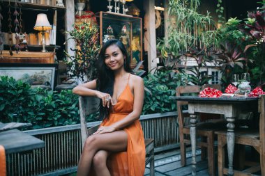 İyimser, genç, formda, uzun saçlı, şık bir yaz elbisesi giyen, ahşap mobilya ve tropikal bitkilerle birlikte açık hava restoranında dinlenen esmer bir kız.