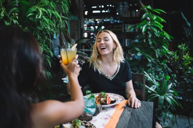 Çok ırklı kadınlar yaz kıyafetleri içinde bardaklarını kokteyllerle tokuştururken tropikal yeşilliklerle dolu egzotik restoranlarda lezzetli yemekler yiyorlar.