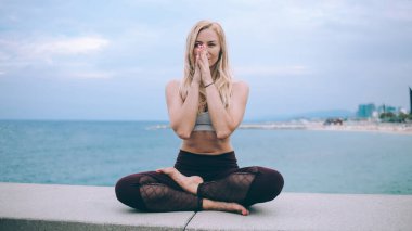 Deniz kenarında meditasyon ve yoga yaparken gülümseyen ve deniz dinleyen mutlu genç bayan bulutlu bir günde kameraya bakıyor.