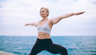 Beyaz kadın yogi Virabhadrasana egzersizi yapıyor. Temiz okyanus havasında antrenman yaparken eşofmanlı zayıf bir kadın, savaşçı pozu veriyor. Vücut uyumu için sağlıklı yaşam tarzından zevk alıyor.