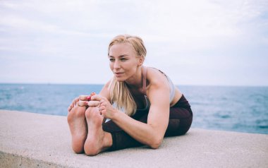 Genç, esnek, çıplak ayaklı, sarışın, siyah taytlı, beyaz bluzlu, yoga pozu veren bir kadın.