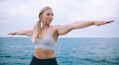 Spor kıyafetli sıska bir kadın yoga antrenmanı sırasında esneme egzersizi yapıyor. Bulutlu bir günde antrenman yaparken başını çeviriyor.